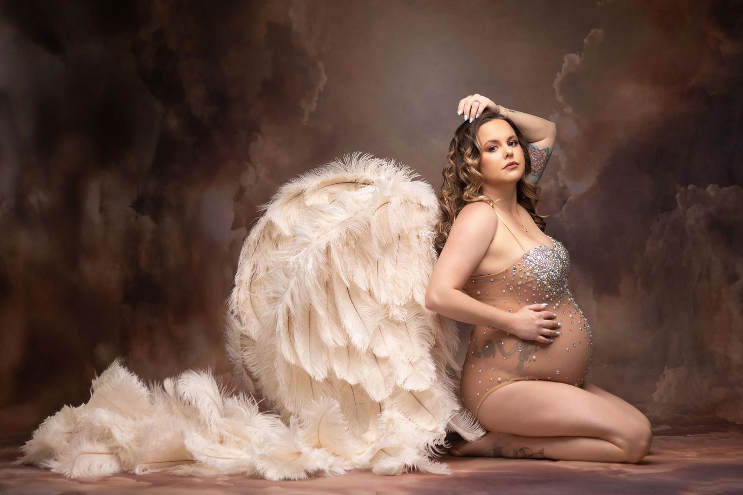 Pregnant woman in jeweled bodysuit poses gracefully with large white feather angel wings against a moody brown studio backdrop.