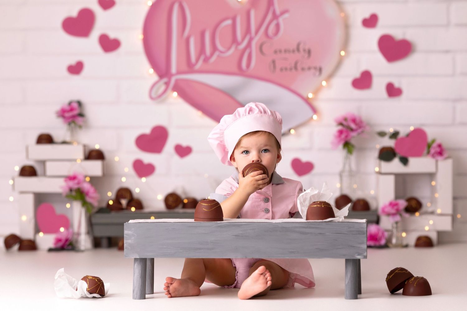 Baby in pink chef outfit sits with chocolates at Lucy's Candy Factory Valentine's Day themed photo shoot.