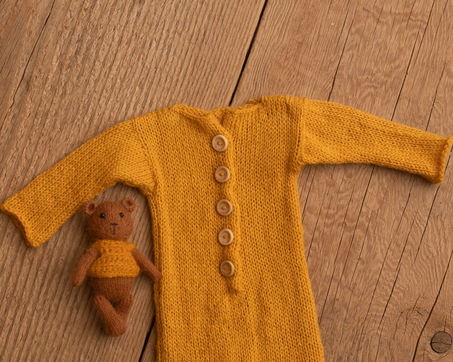 Mustard yellow knitted baby romper with wooden buttons beside a small brown teddy bear on a wooden floor.