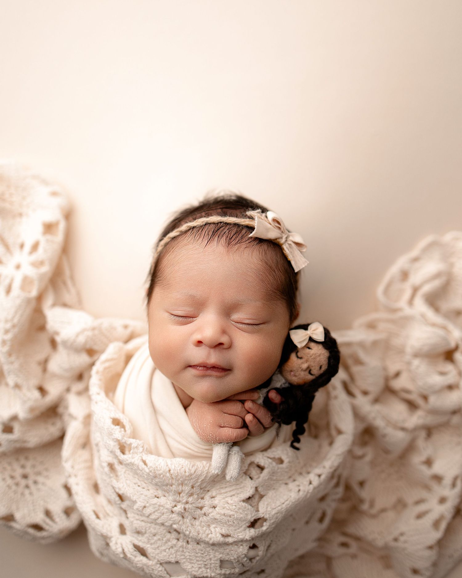 Sleeping newborn baby wrapped in cream lace swaddle, wearing bow headband, holding a small doll.