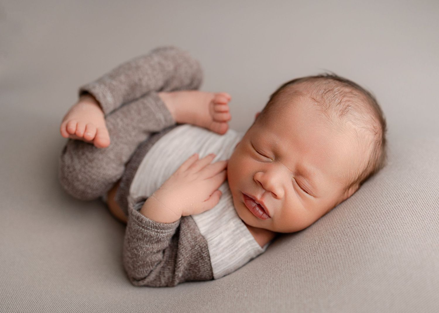 A peaceful newborn baby sleeps in a gray knit outfit on a soft gray backdrop.