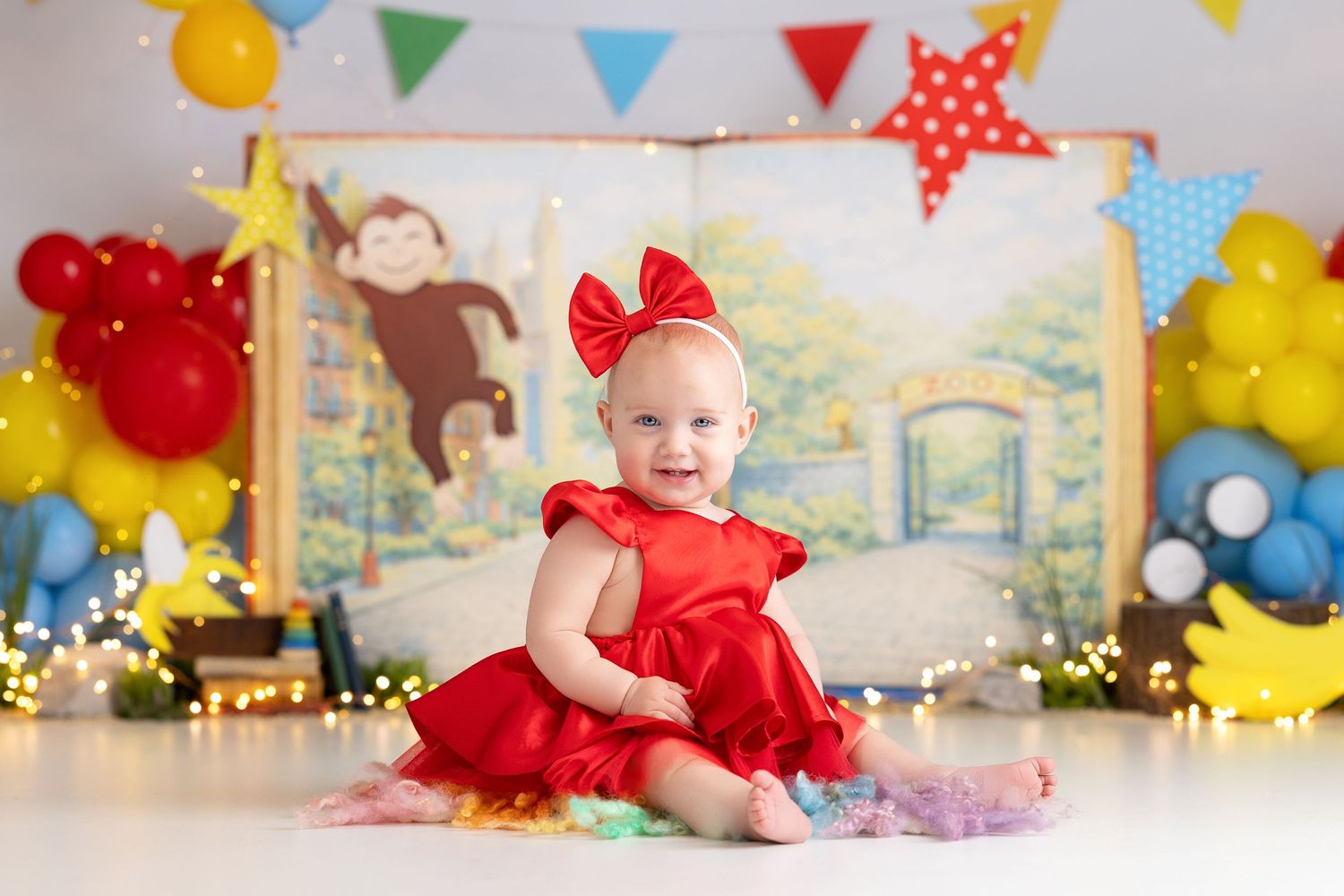 Baby girl in red dress sits smiling at Curious George themed first birthday cake smash photo session.