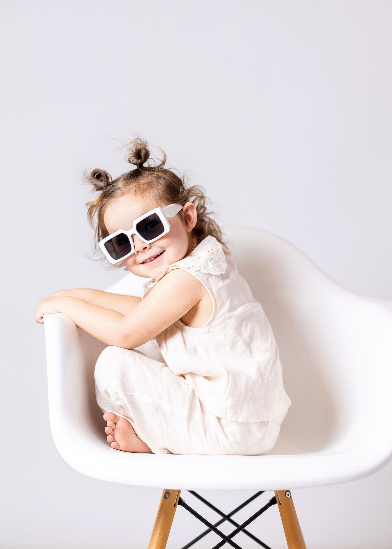 Cute little girl in white dress and sunglasses sitting cross-legged on a modern white Eames chair, smiling.
