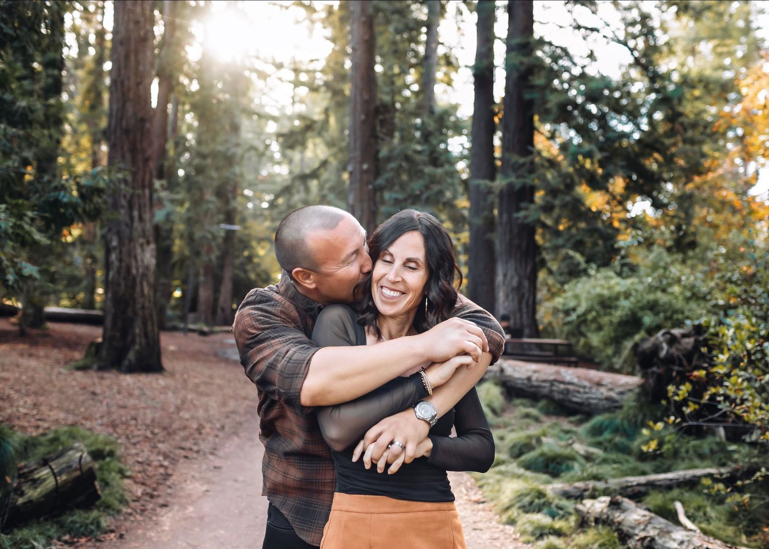 Couple embracing and laughing together on a forest path surrounded by tall redwood trees.