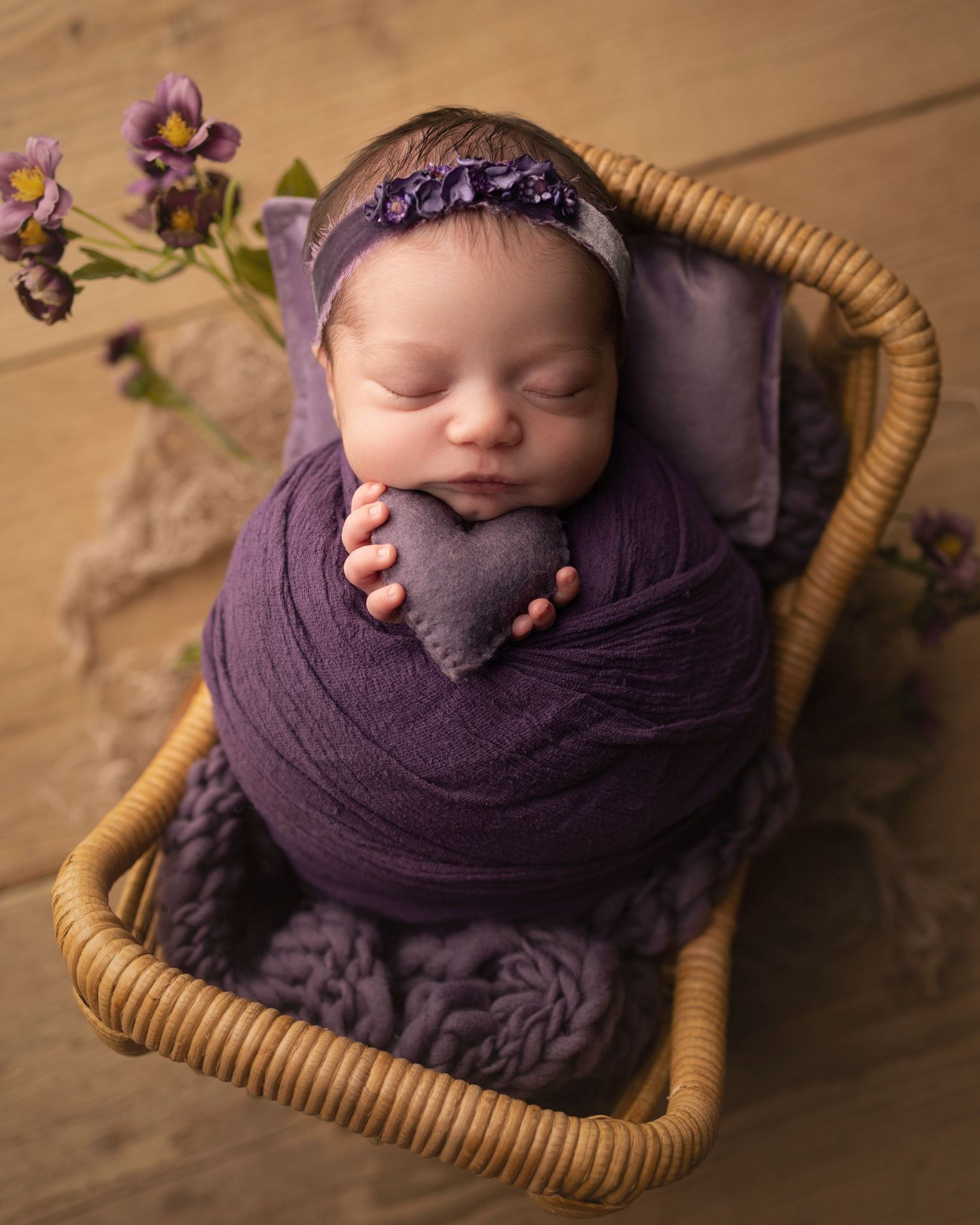 Newborn baby girl wrapped in purple fabric holding a heart prop in a basket, professional studio photography in Vacaville California