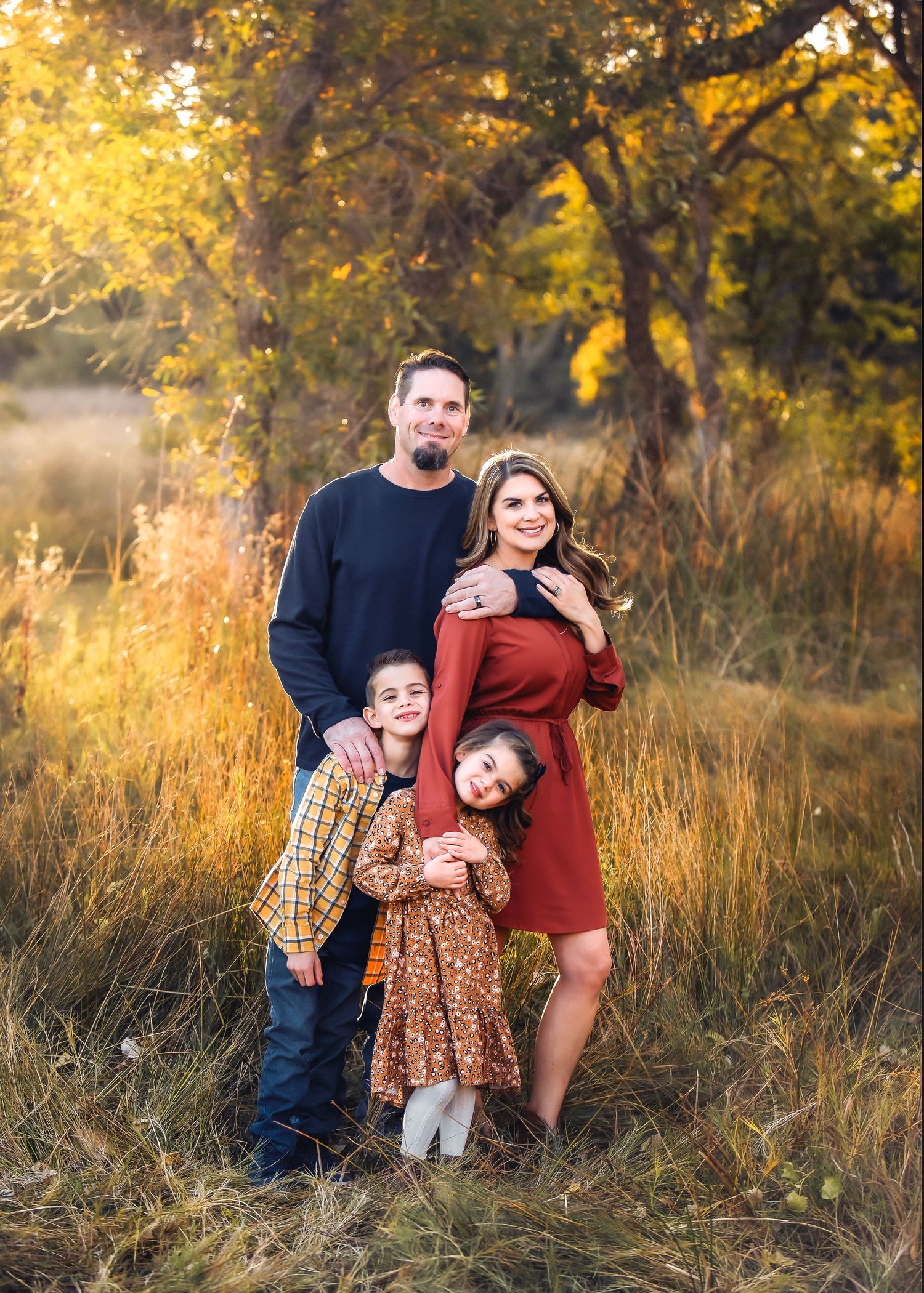 Family of four poses in golden autumn field, parents and two children smiling warmly in coordinated fall outfits.