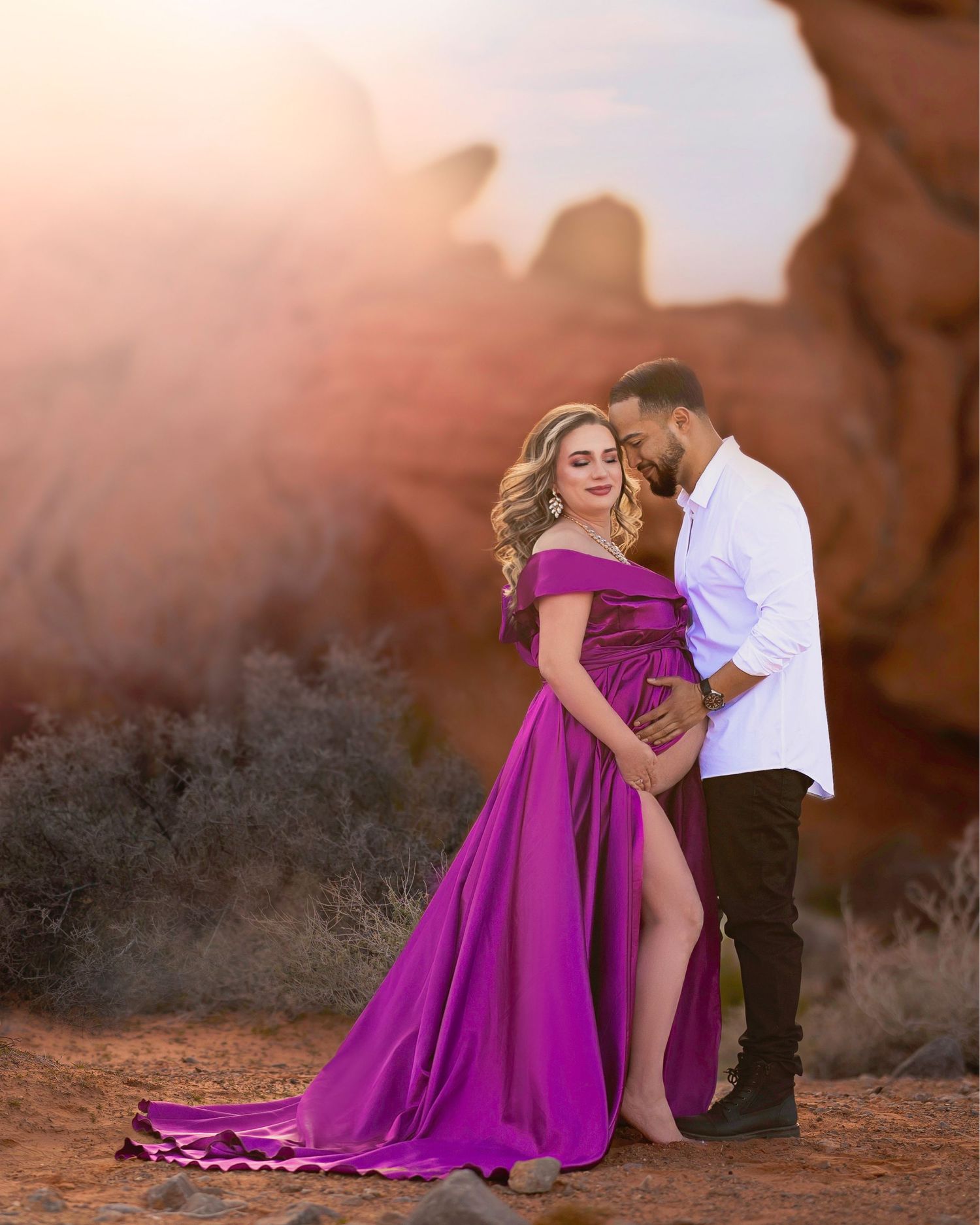 Couple in desert maternity photoshoot, woman in purple off-shoulder gown, man in white shirt, red rock backdrop.