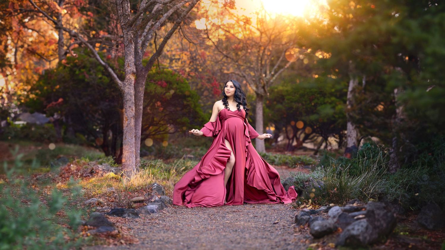 A woman in a flowing red gown poses gracefully on a garden path surrounded by autumn trees at golden hour.