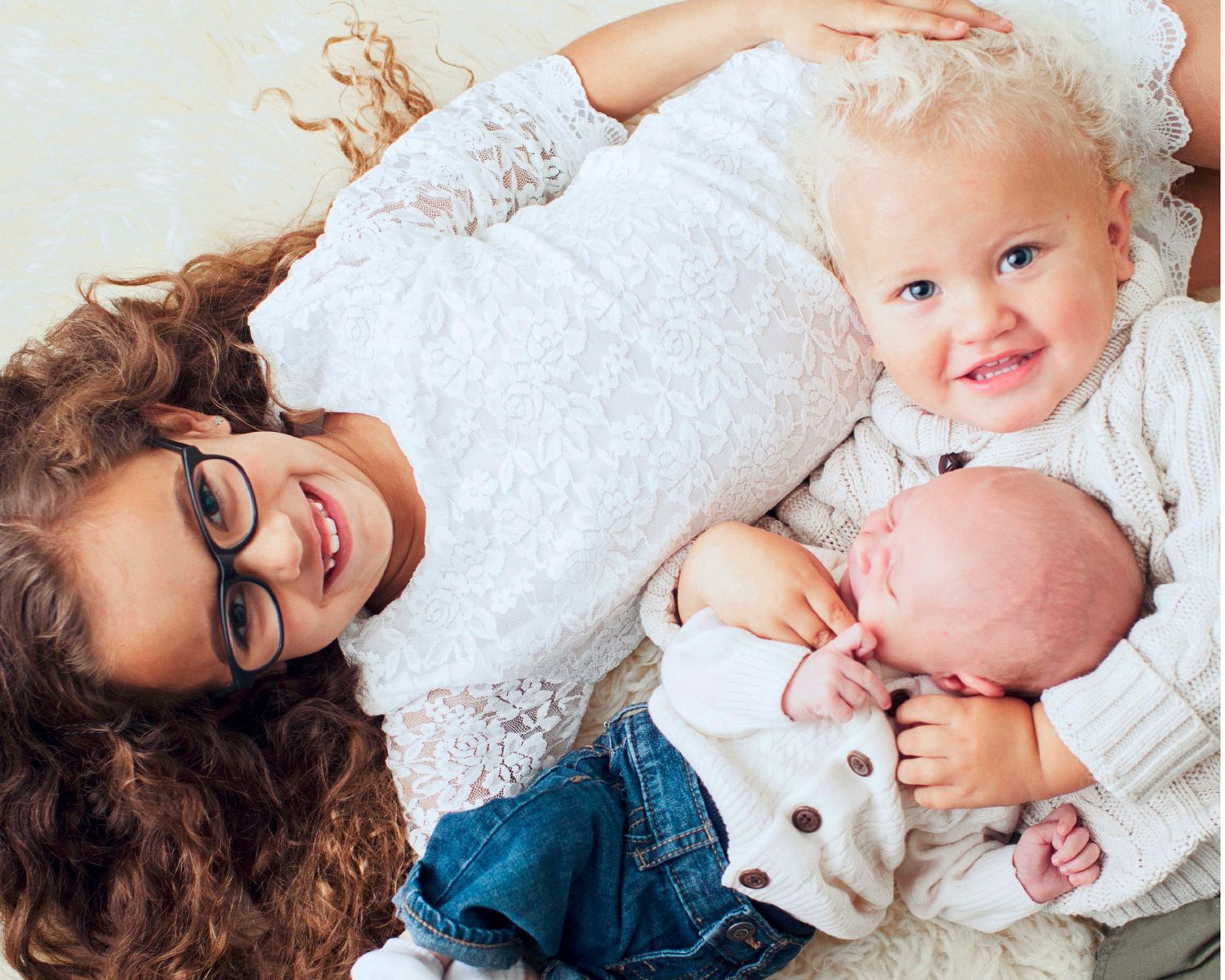 Mother with curly hair and glasses lying down smiling with toddler and newborn baby in white outfits.