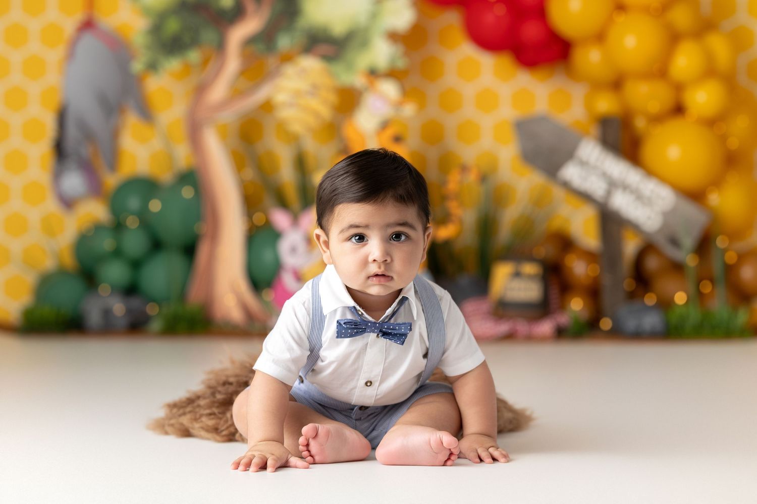 winnie the pooh theme, Baby boy in bow tie sits on white floor before colorful balloon backdrop at first birthday cake smash.