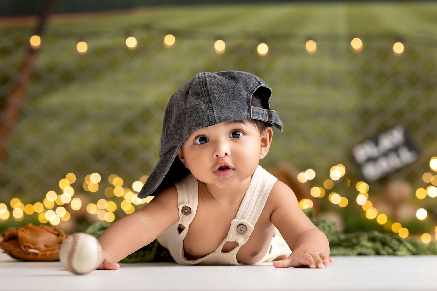 Adorable baby in denim cap and suspenders poses with baseball among bokeh lights in a sports-themed photo shoot.