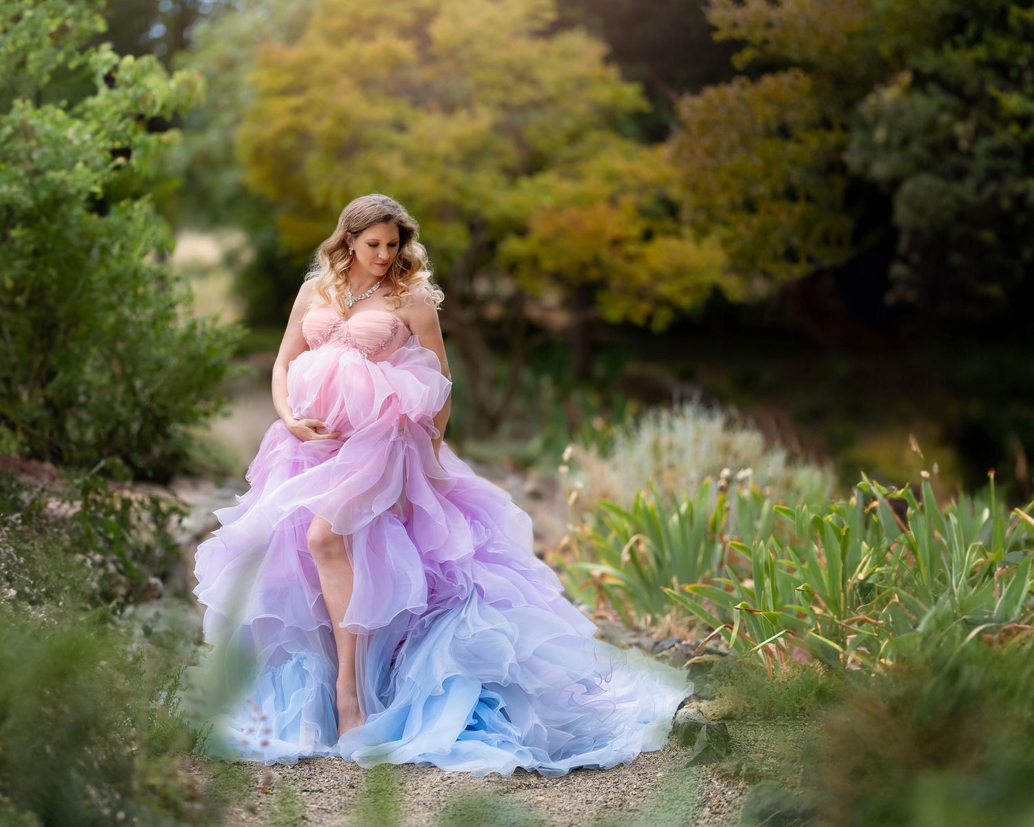 Woman in colorful rainbow tulle maternity gown posing outdoors in a lush garden setting.