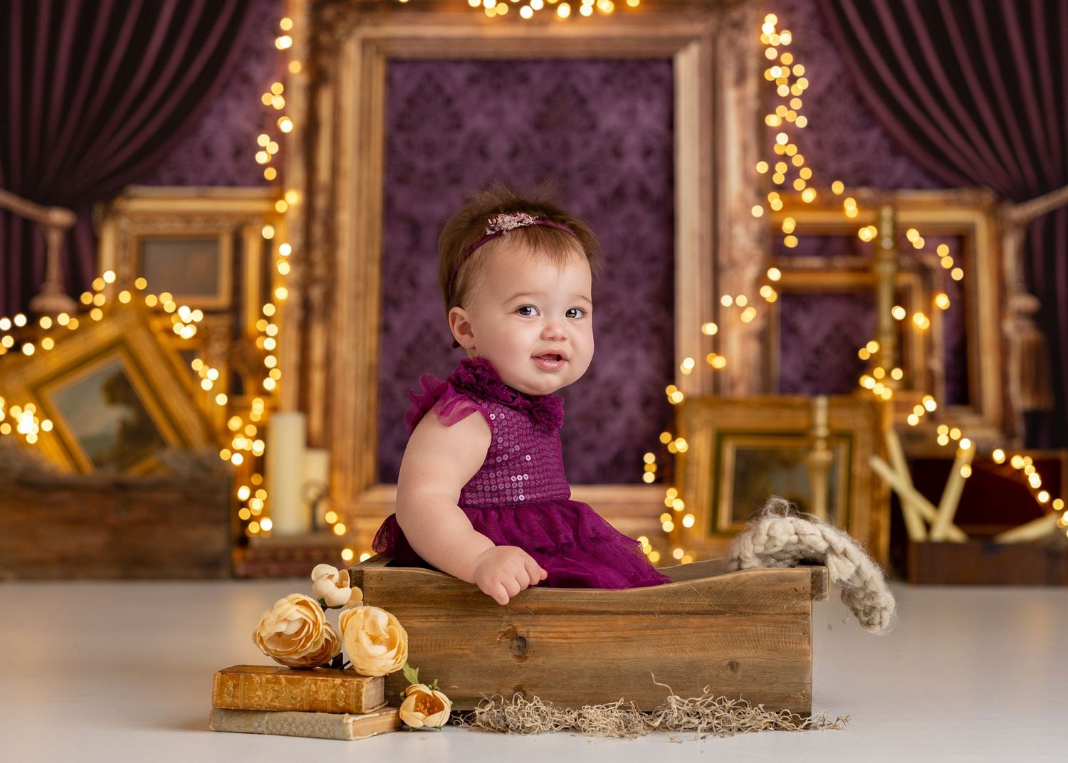 Baby girl in purple dress sits in wooden box surrounded by gold frames and fairy lights in elegant portrait, Bridgerton theme .