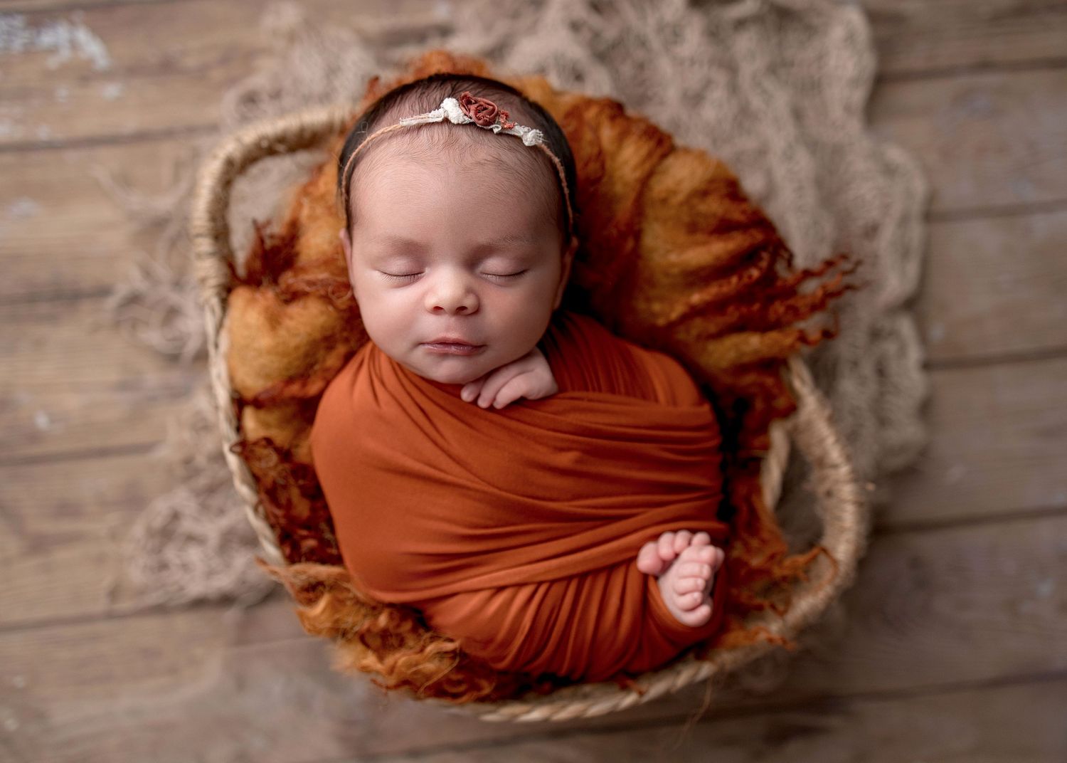 Newborn baby girl wrapped in rust orange swaddle sleeping peacefully in a woven basket with floral headband.