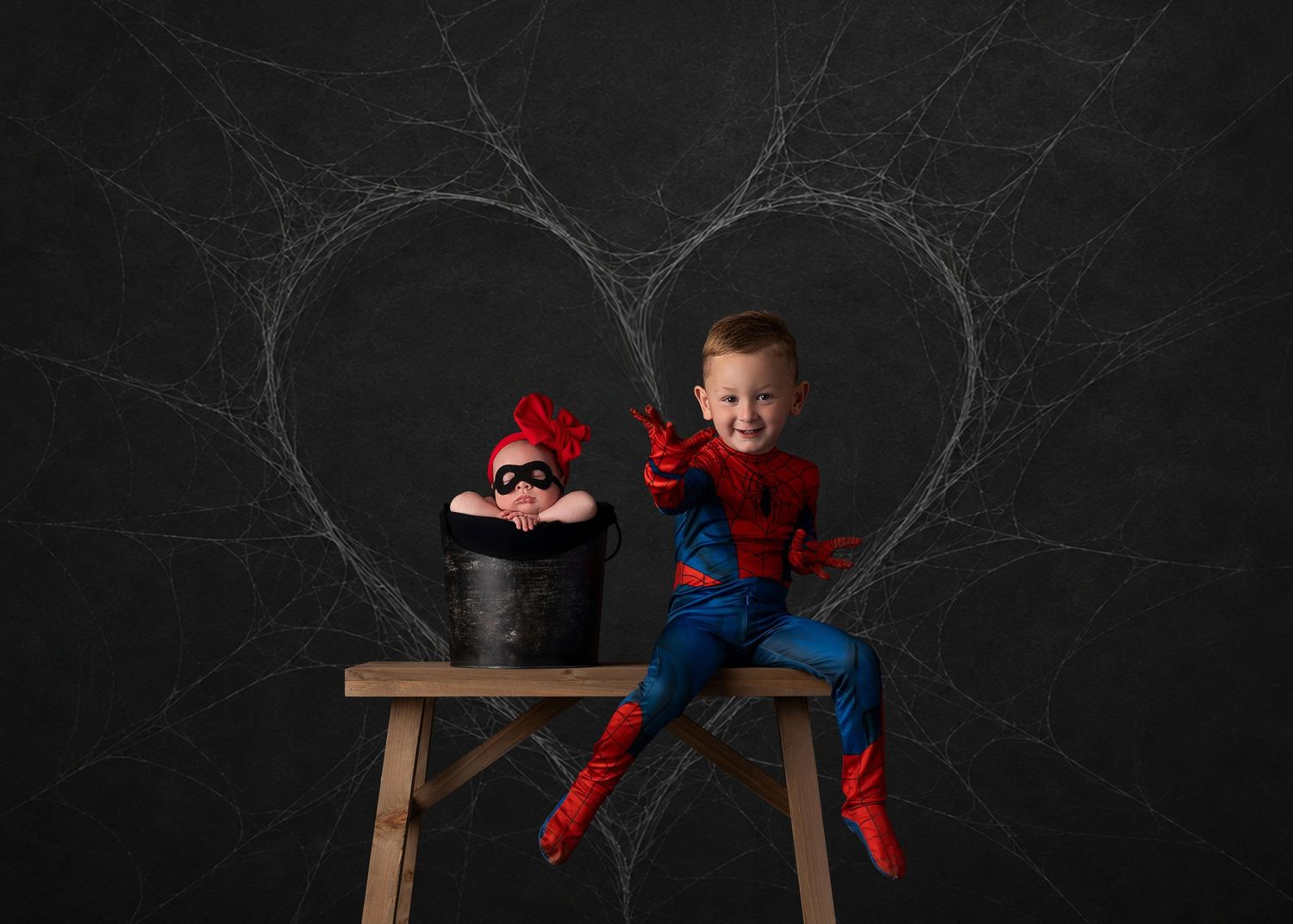 Boy in Spiderman costume sits beside baby in black bucket, both posed against dark heart web backdrop.