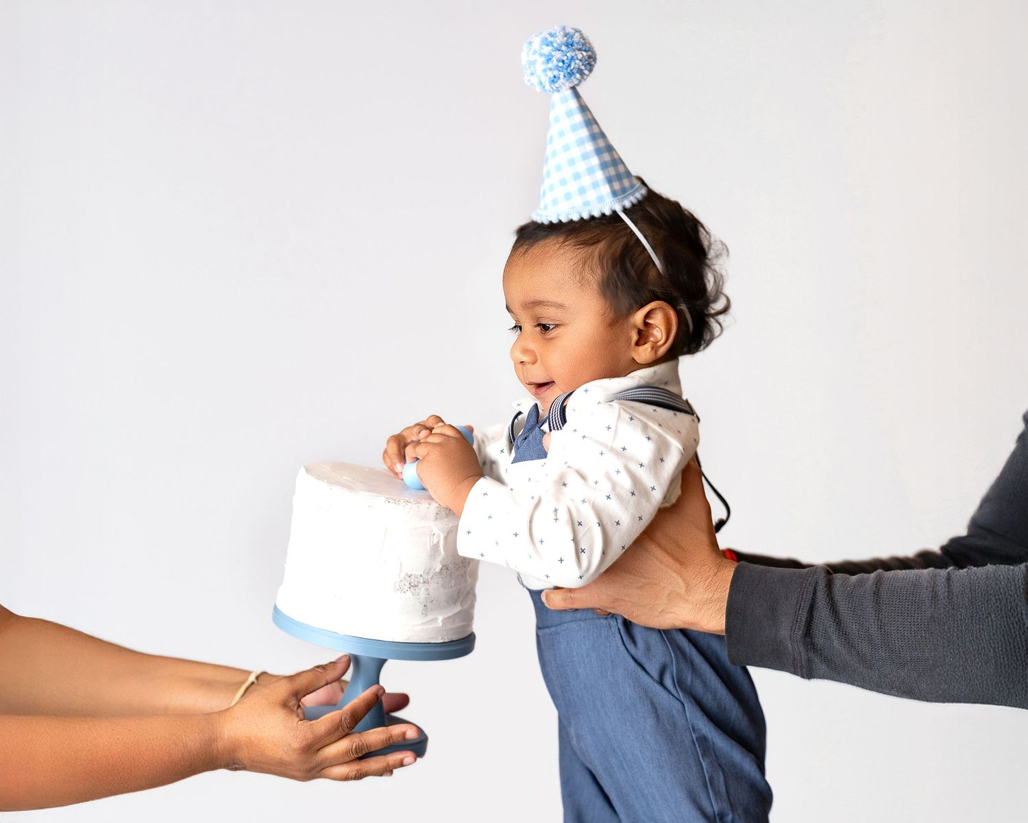 Toddler in blue party hat smiling at white birthday cake held by adults on gray background.