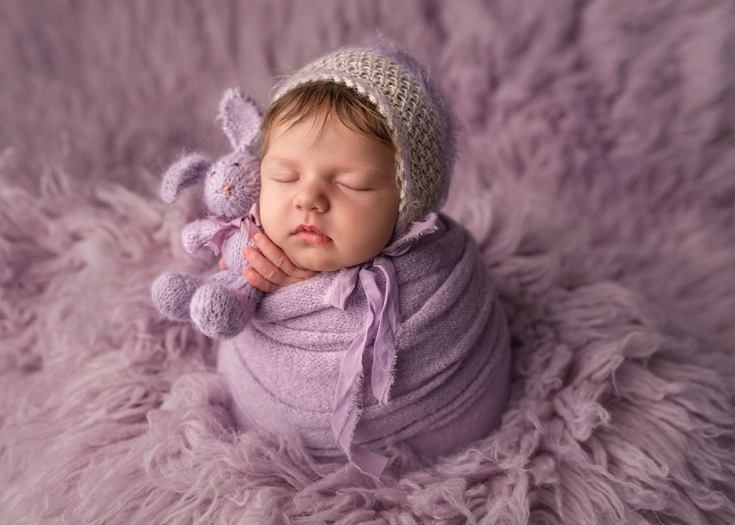 Sleeping newborn baby wrapped in purple knit blanket wearing gray bonnet on lavender fur backdrop.
