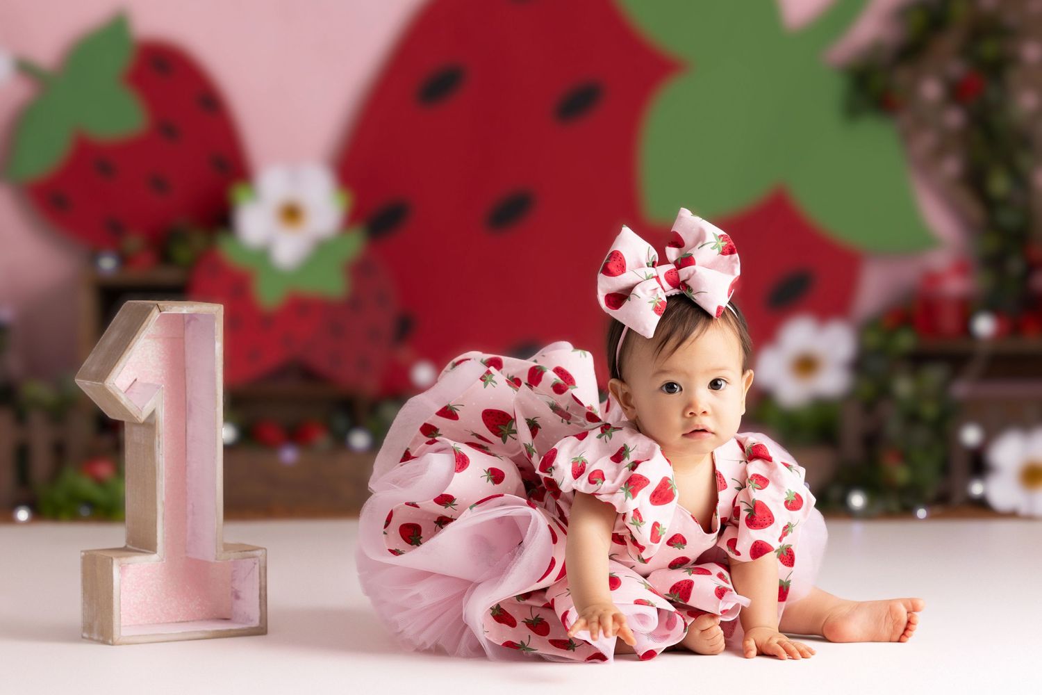 Baby girl in strawberry dress with bow headband sits beside a number 1 block at a first birthday photo shoot.