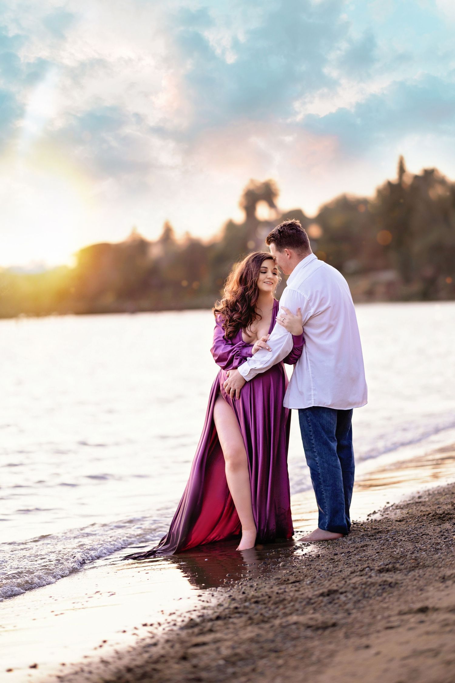 Couple embracing on a beach at sunset, woman in purple flowing gown, man in white shirt and jeans.