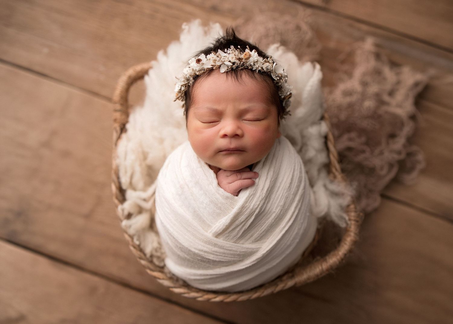 Sleeping newborn baby wrapped in white swaddle with floral crown in wicker basket on wood floor.
