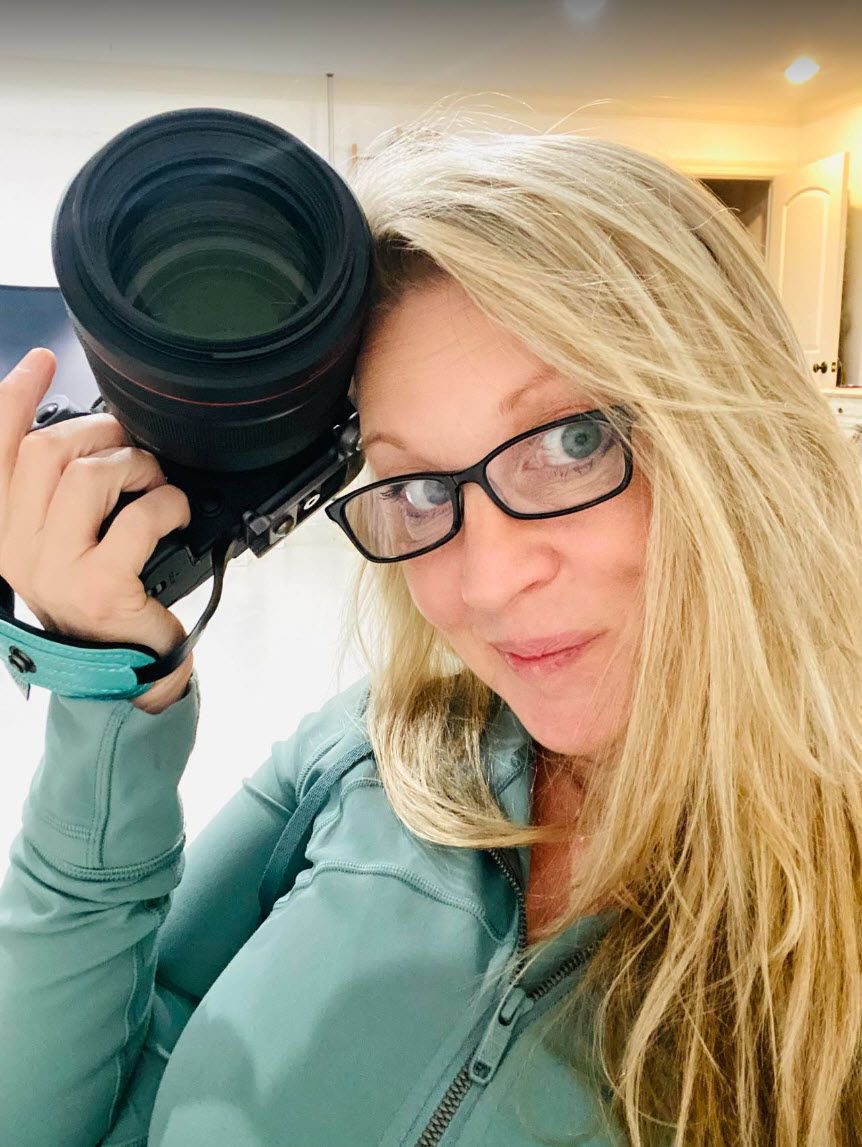 A blonde woman with glasses holds a professional camera close to her face indoors.