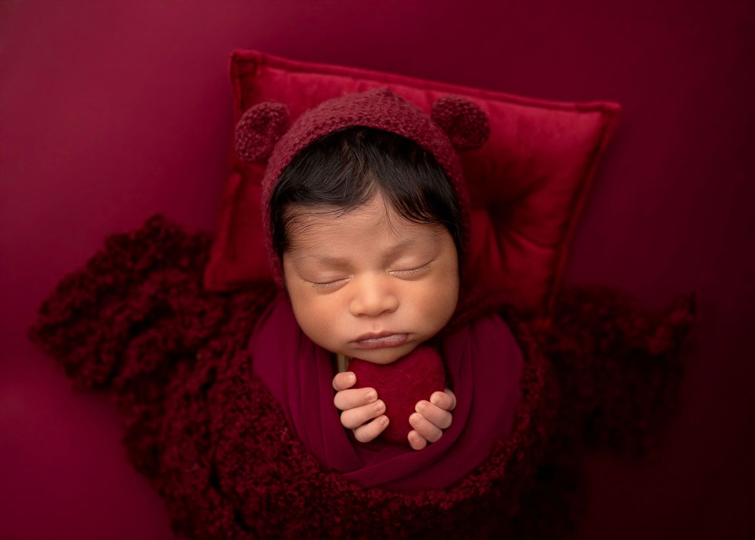 Newborn baby sleeping peacefully wrapped in burgundy swaddle with red bear bonnet on matching velvet background.