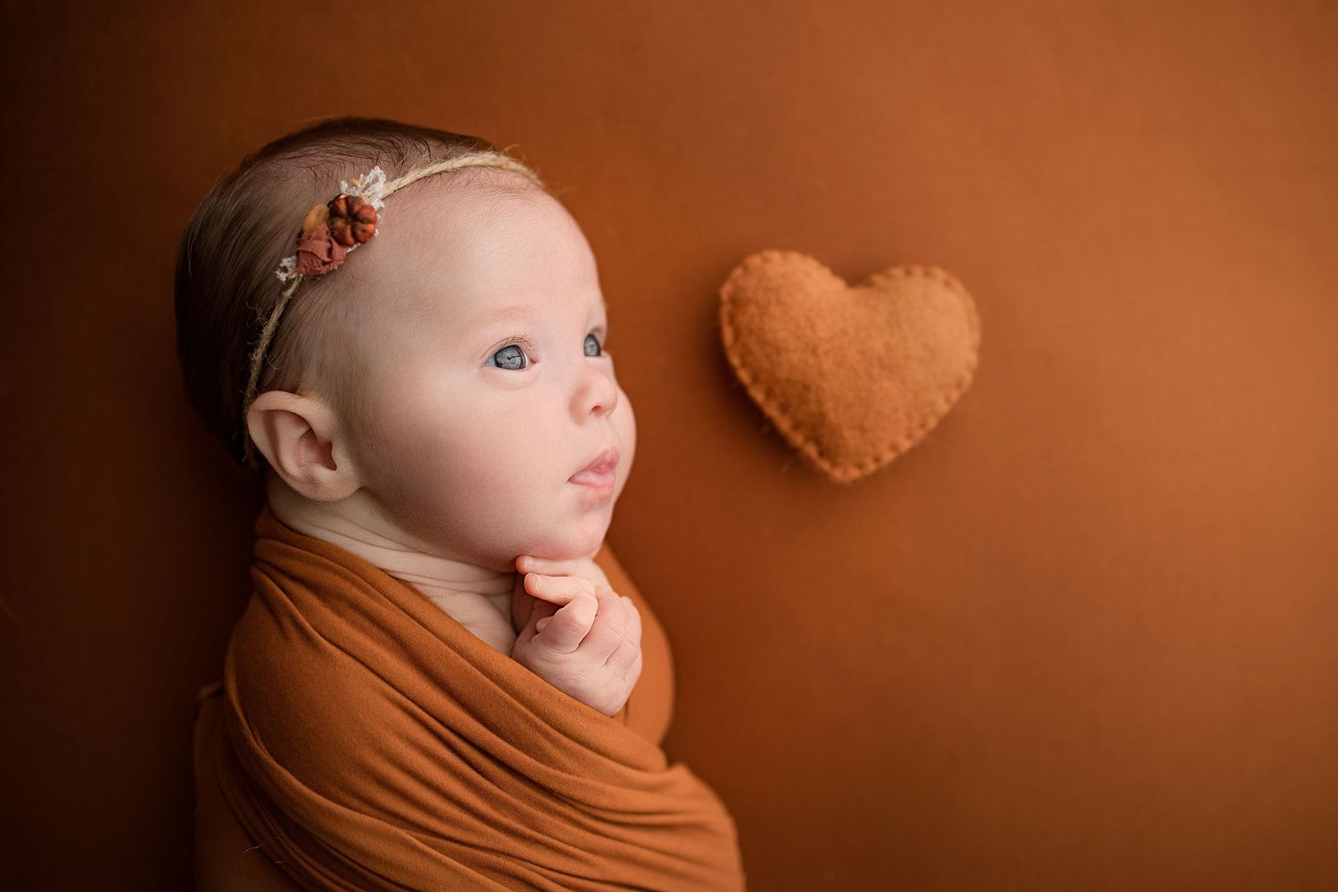 A newborn baby wrapped in a burnt orange swaddle lies next to a felt heart on a matching background.