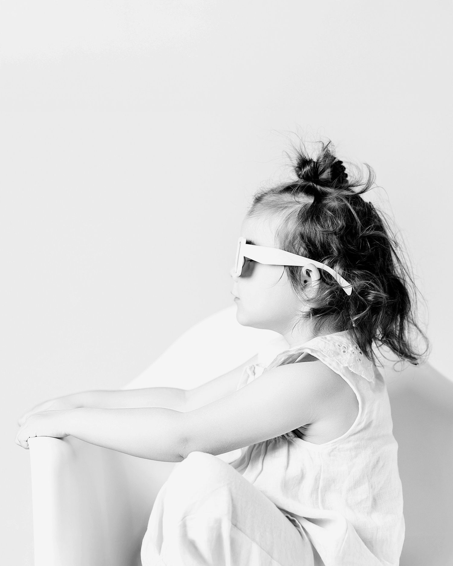 Black and white photo of a young girl with a bun hairstyle wearing sunglasses, sitting and looking sideways.