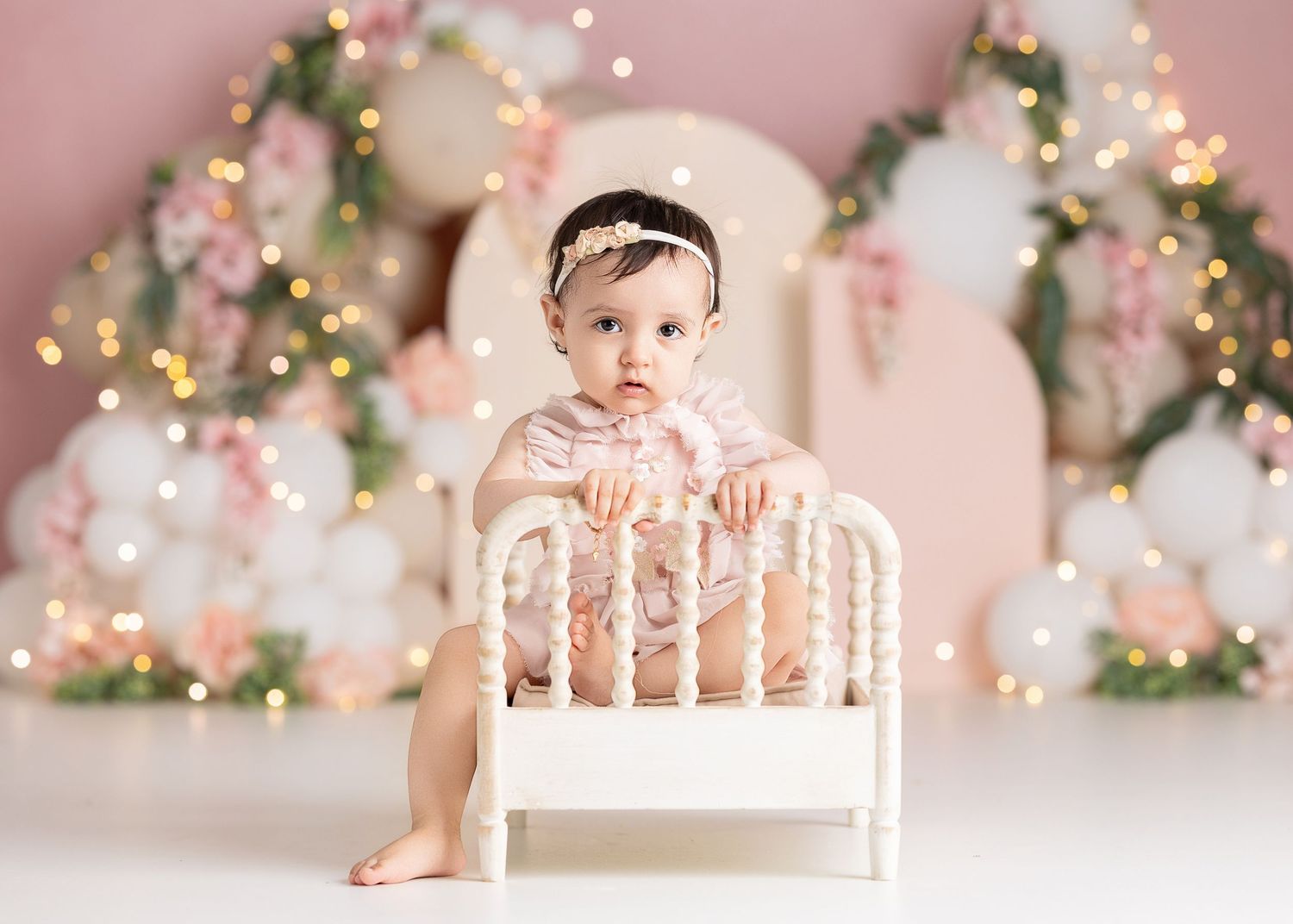 Baby girl in pink outfit sits on white mini bed with balloon arch and fairy lights backdrop.