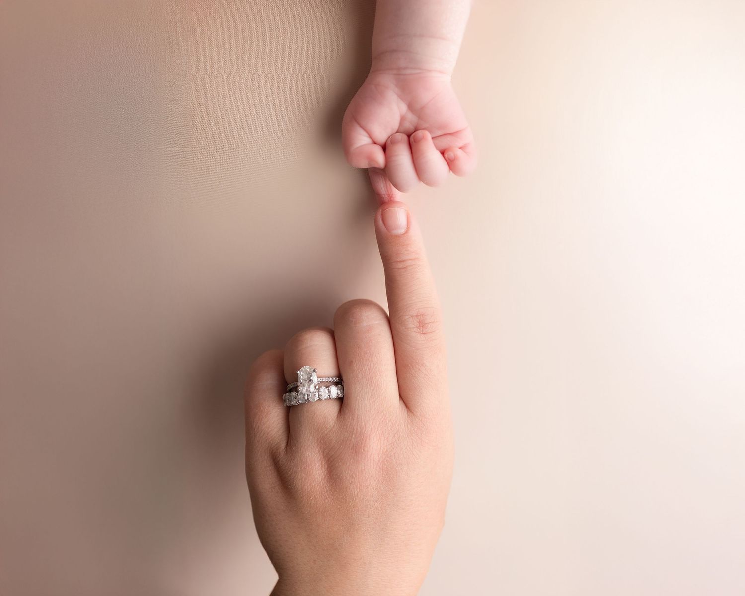 A newborn baby grasping an adult's finger adorned with a diamond engagement and wedding ring stack.