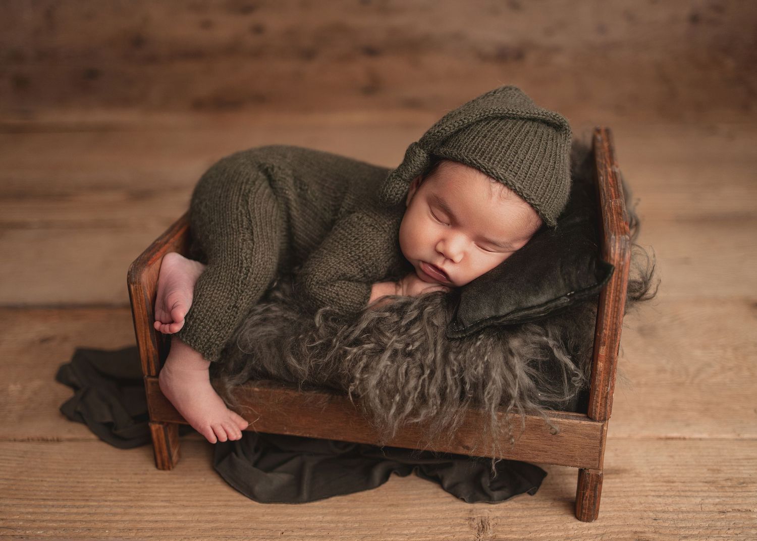 Sleeping newborn baby in olive green knit outfit and hat posed in a small wooden bed with fur blanket.