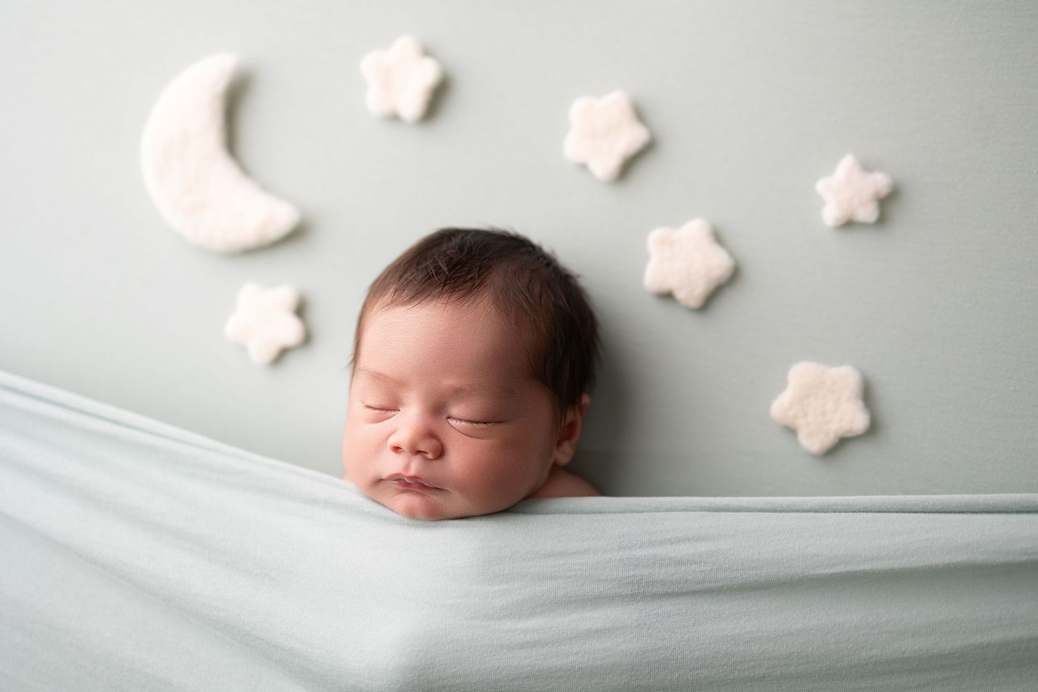 Peaceful newborn baby sleeping wrapped in soft blue fabric with white felt moon and stars decorations on mint wall.