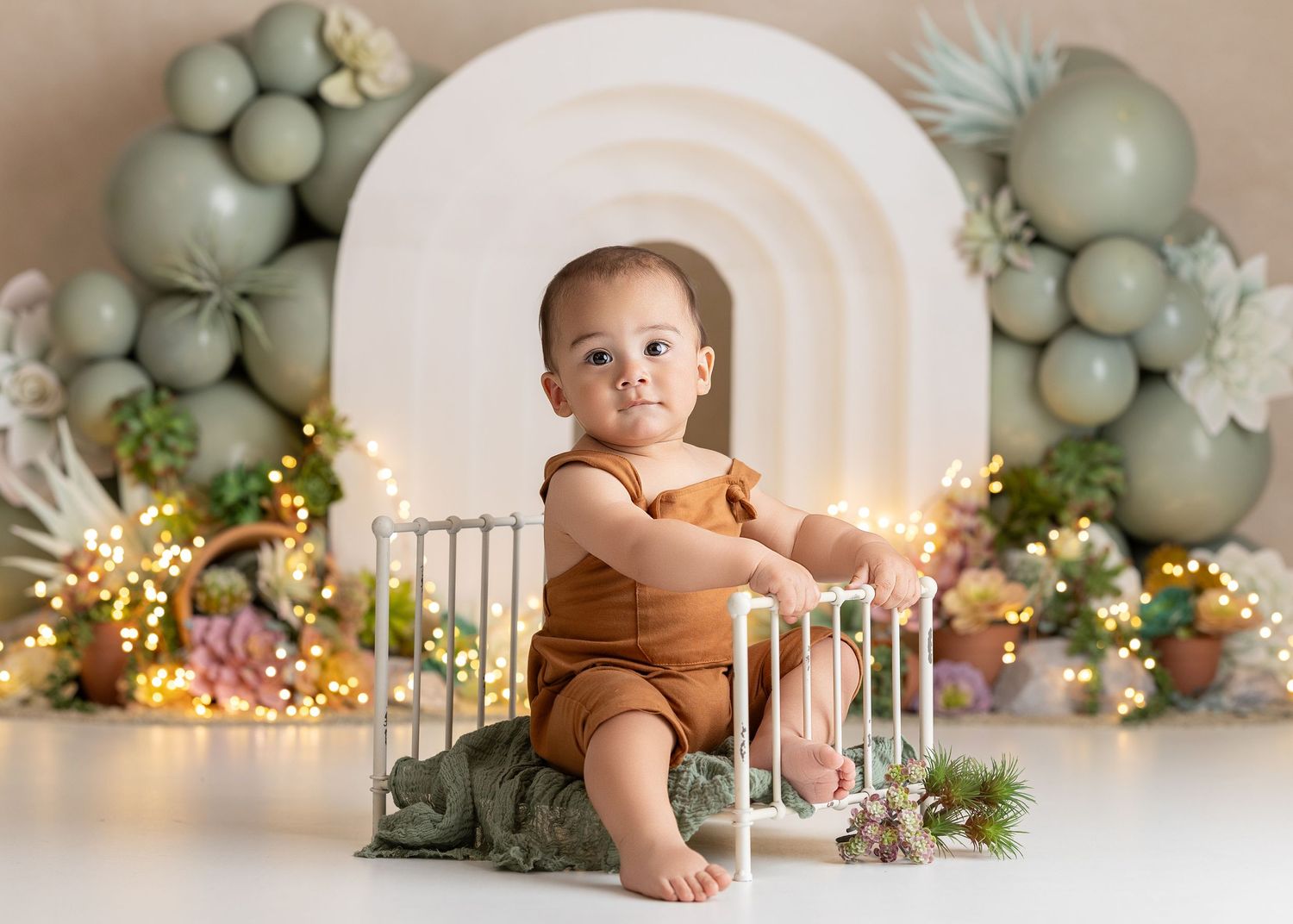 Adorable baby in rust romper sits in mini crib with sage green, succulents, balloon arch and fairy lights backdrop.