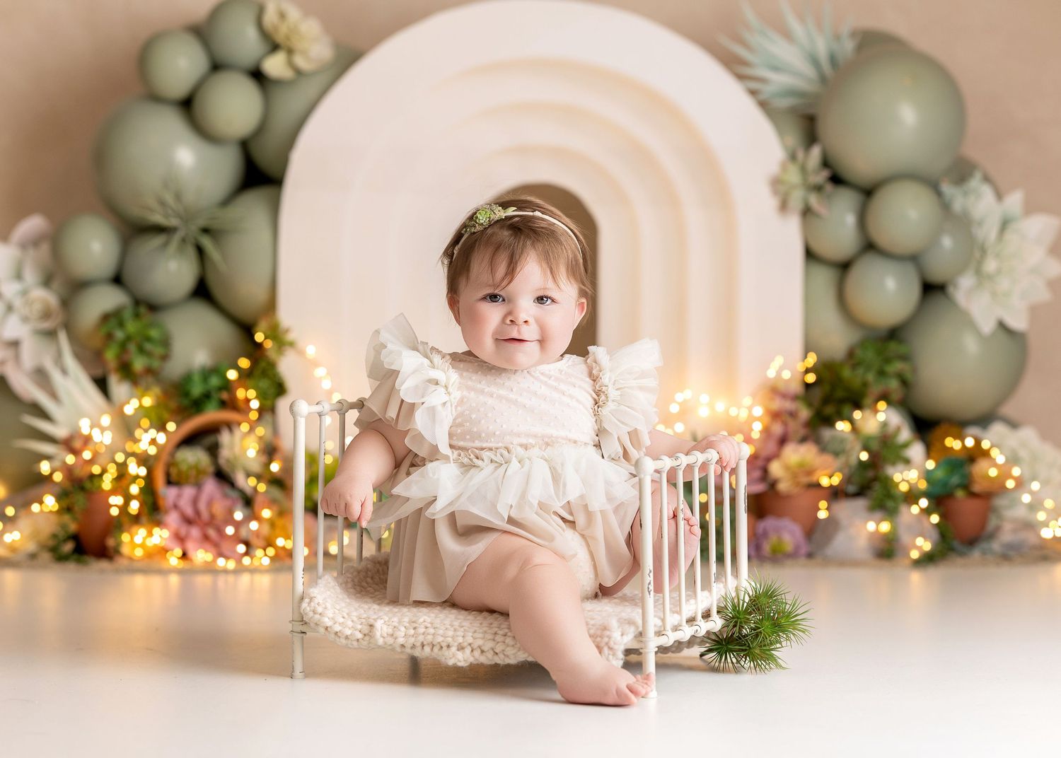 Baby girl in cream dress sits on mini white bed with sage green balloon arch, Succulents and fairy lights backdrop.