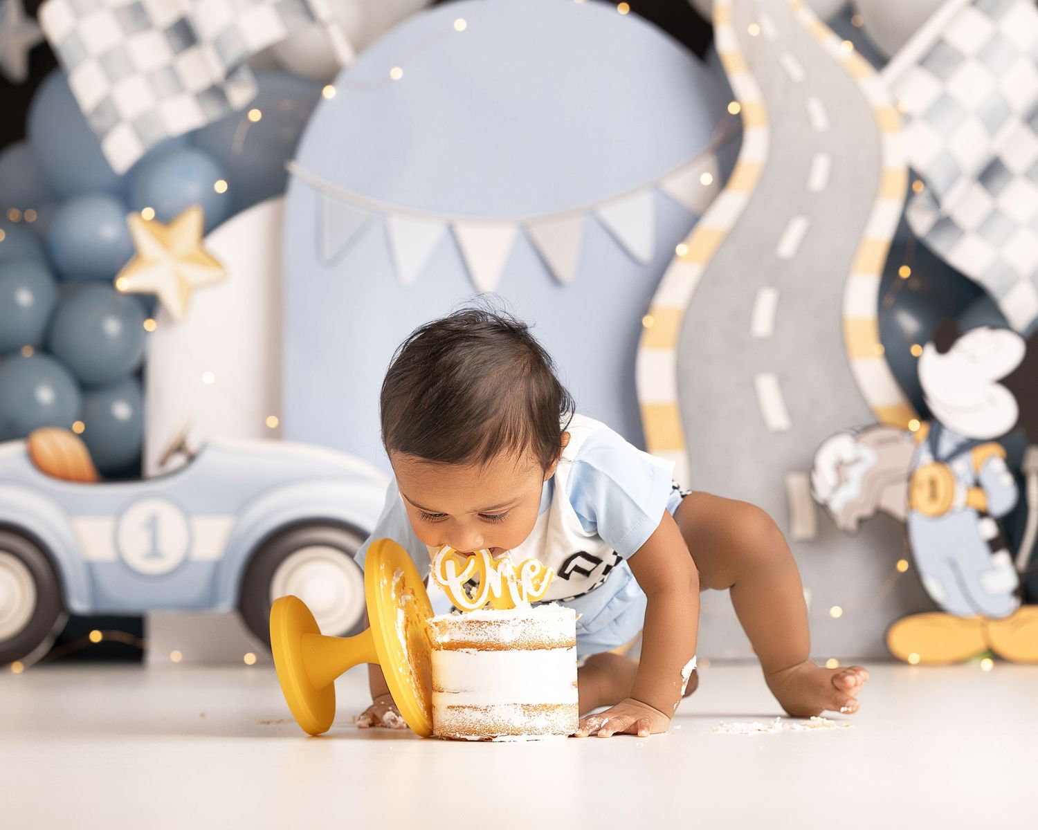 Baby smashing first birthday cake at a race car themed photo shoot with balloons and decorations.