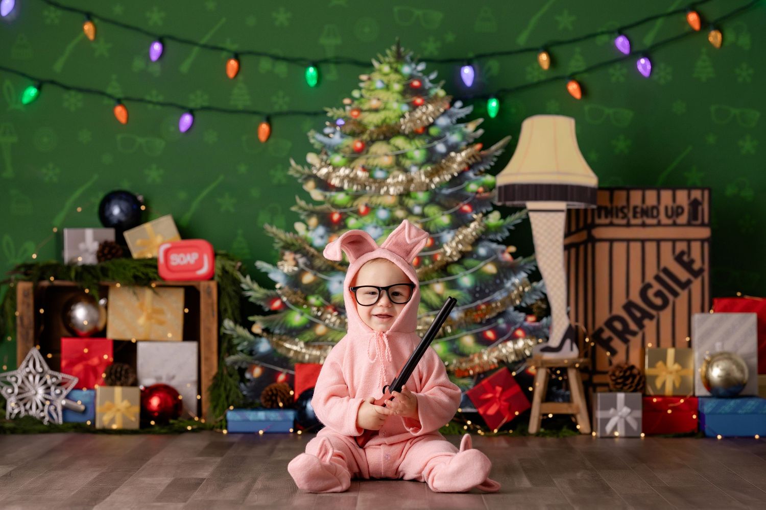Baby in pink bunny suit with glasses poses by decorated Christmas tree with gifts and fragile leg lamp prop, Christmas Story theme.