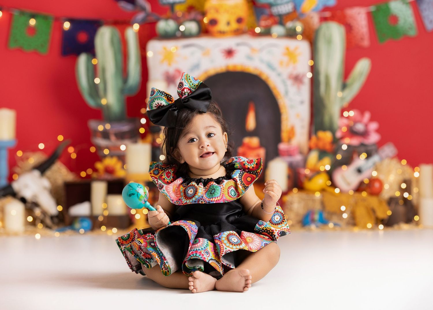 Smiling baby girl in colorful dress with bow sits before festive Mexican-themed backdrop with cactus and fairy lights.
