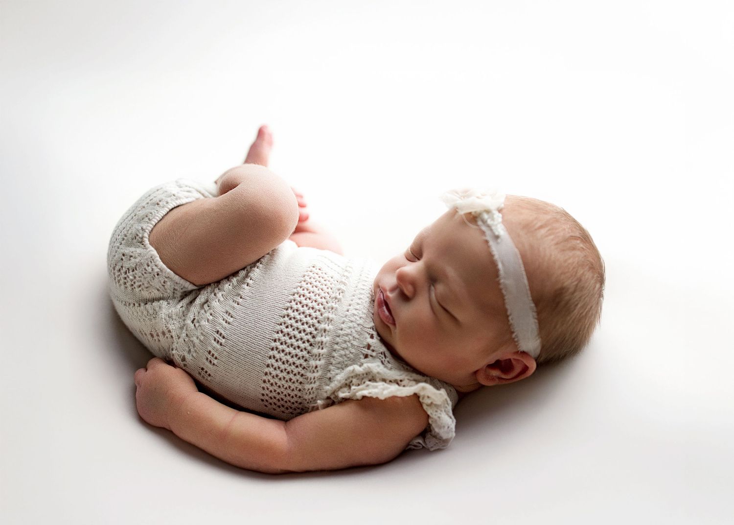 Sleeping newborn baby in white knit romper and headband posed curled up on white background.