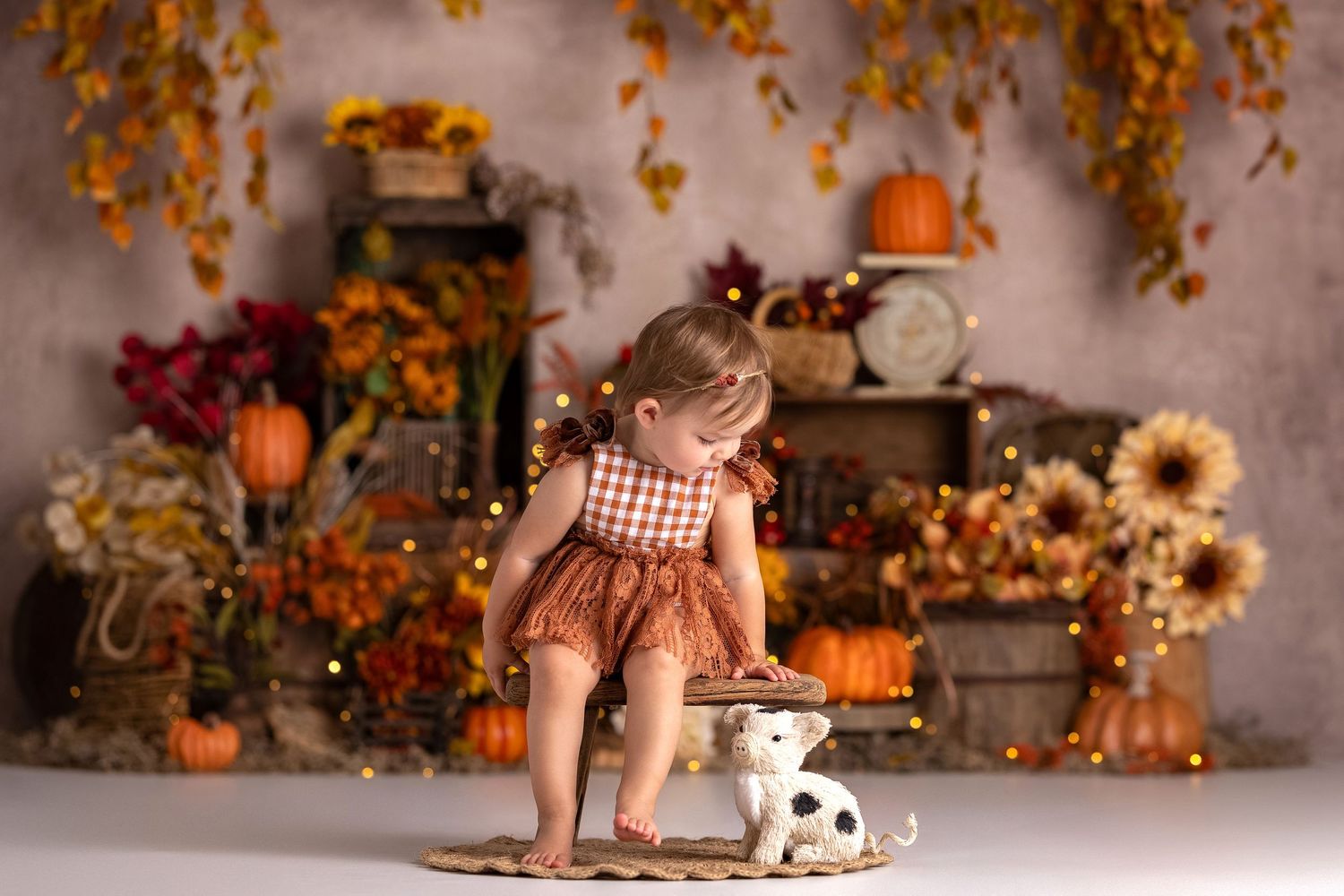 Toddler in fall outfit with plaid top and brown skirt poses near pumpkins, sunflowers, and a toy dog.