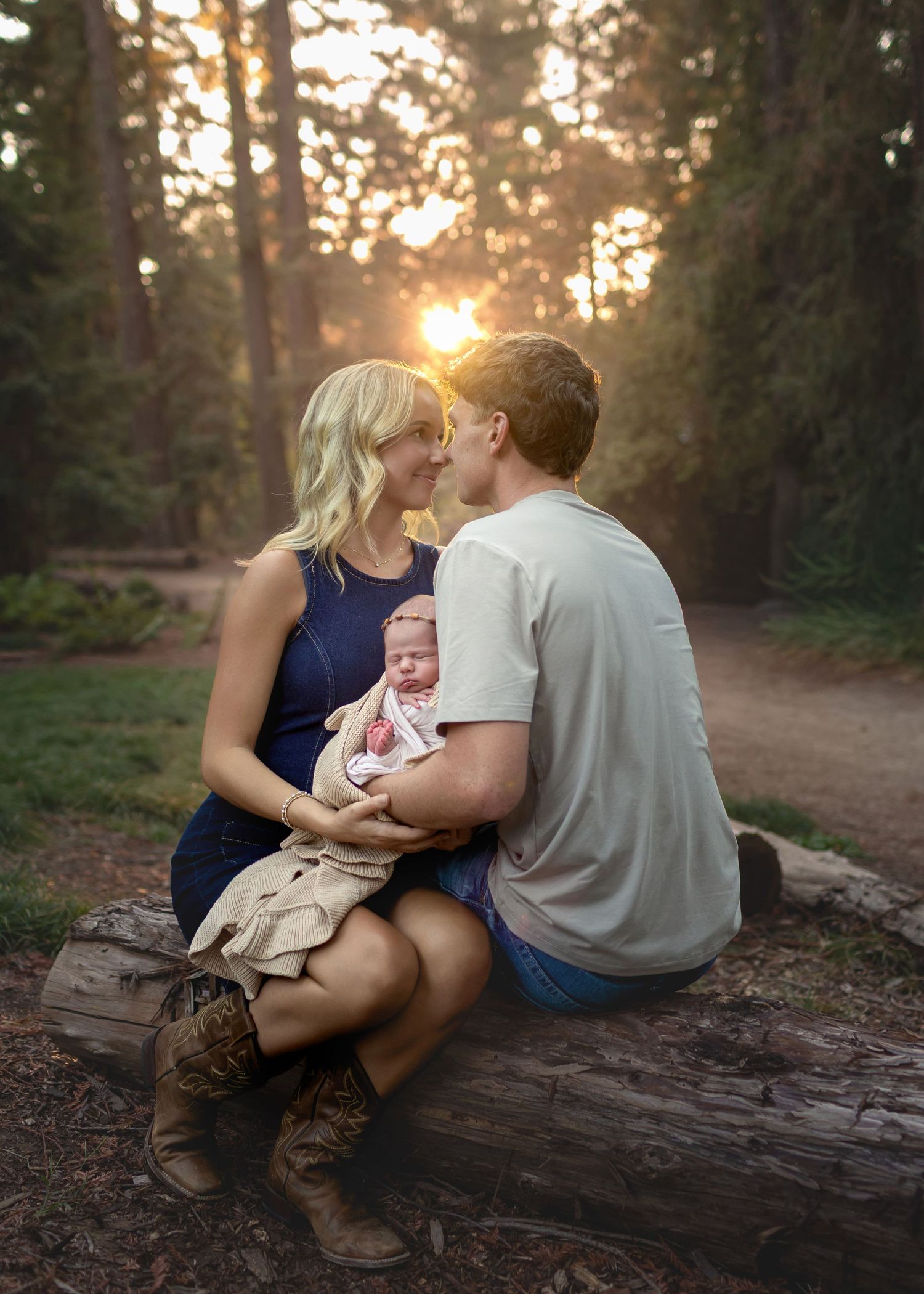 Couple sitting on a log in a sunlit forest at sunset, gently holding their newborn baby while leaning in for a tender moment.