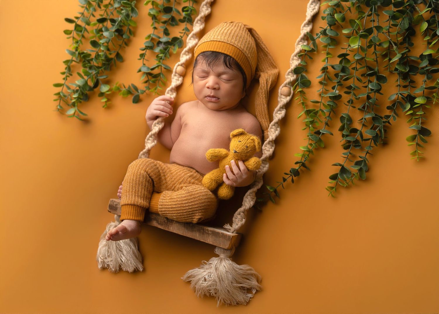 Newborn baby in mustard knit outfit sleeping on a tiny wooden swing with a teddy bear and green vine backdrop.