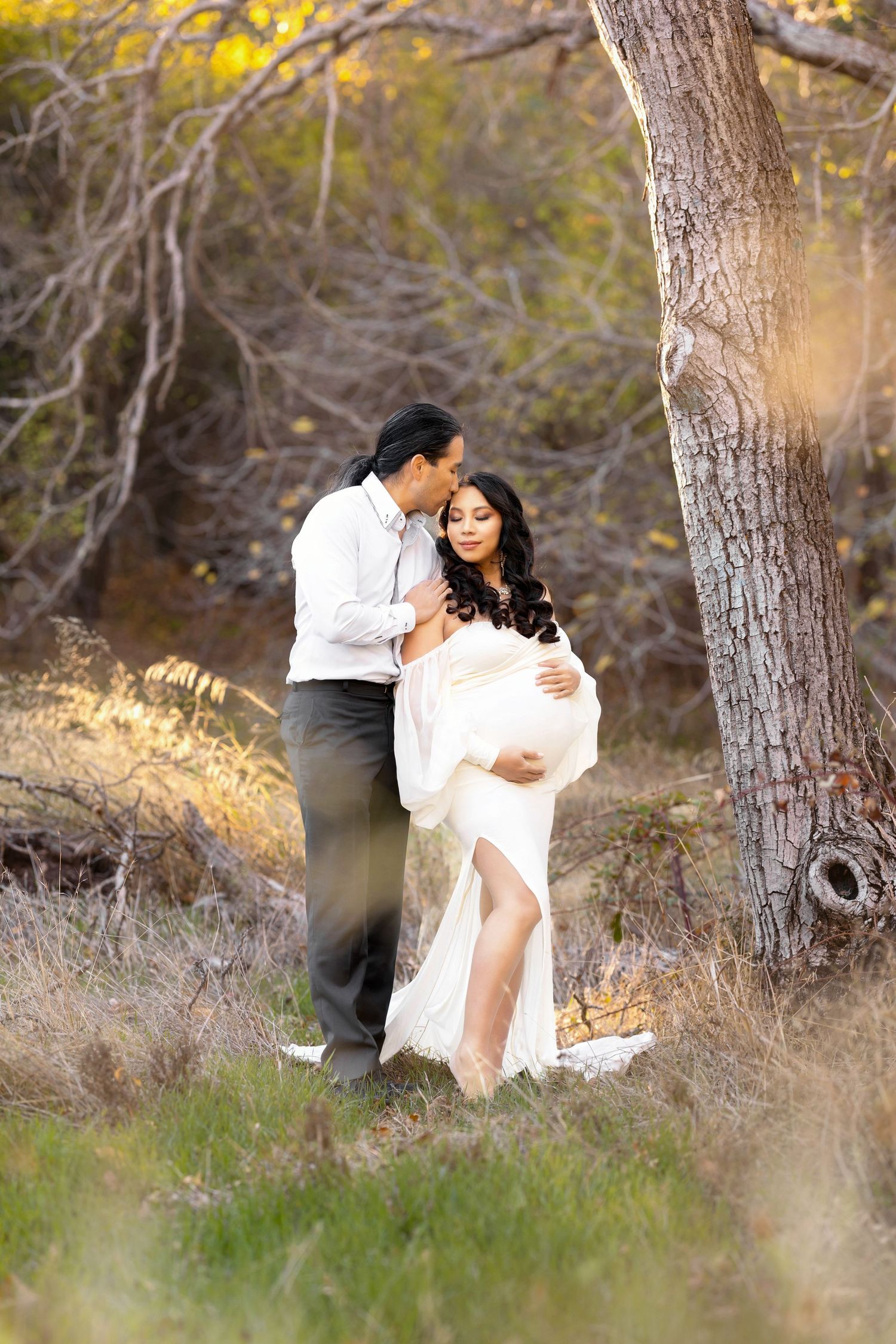 Loving couple shares a tender moment near a tree, with pregnant woman in a white flowing gown outdoors.