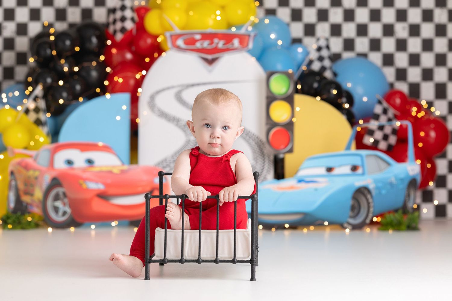 Baby in red dress sits on mini bed with Cars movie themed backdrop featuring Lightning McQueen and balloons.