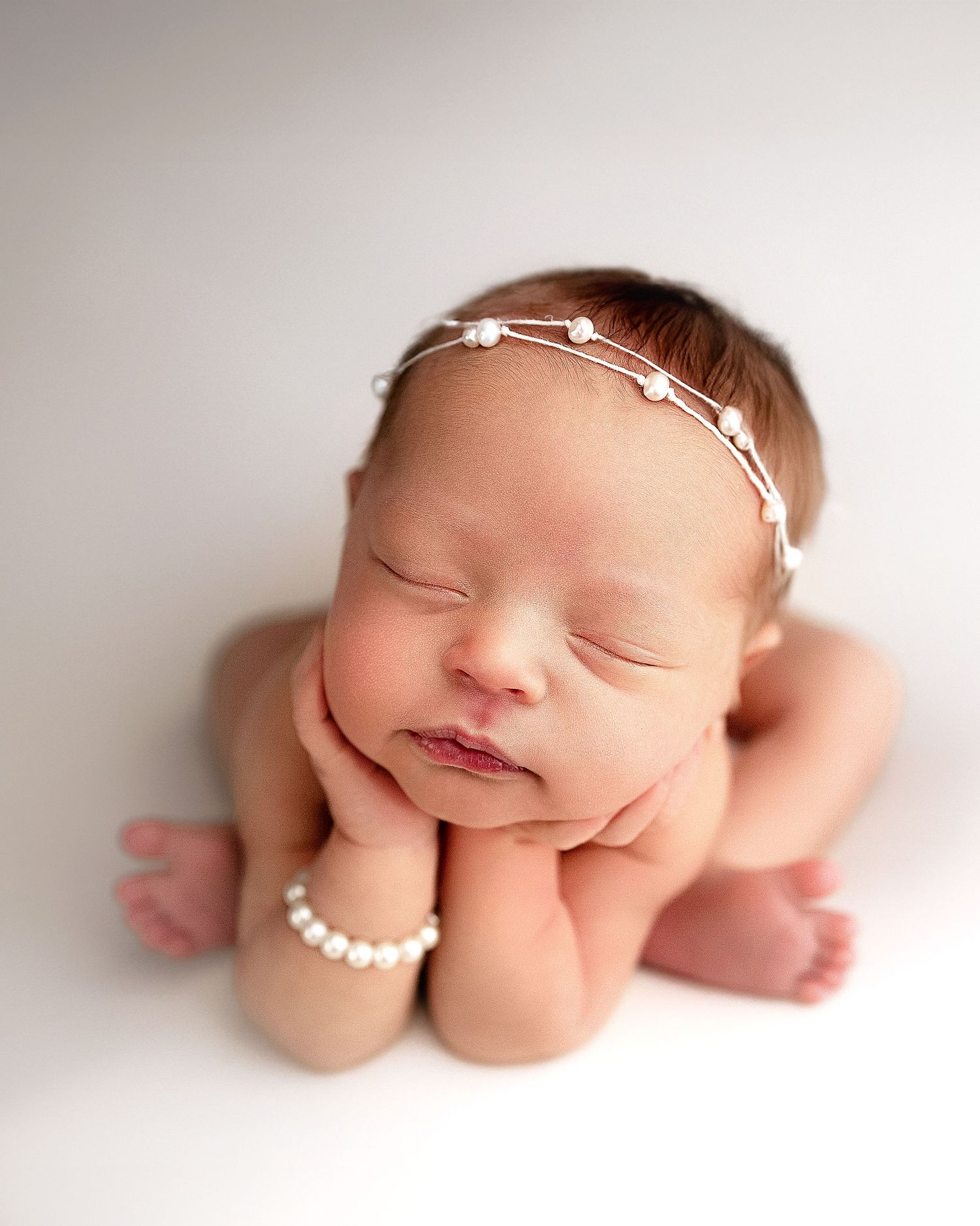 Newborn baby girl in froggy pose on a white backdrop with pearl headband and bracelet, studio photography in Vacaville Californi