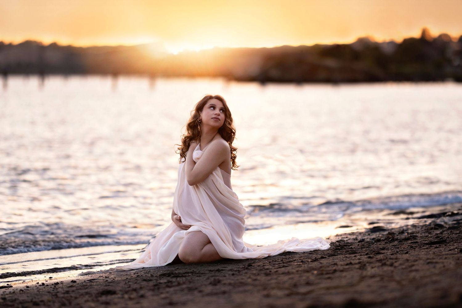 Young woman in white dress sitting on beach shore at golden sunset, arms crossed, gazing upward dreamily.