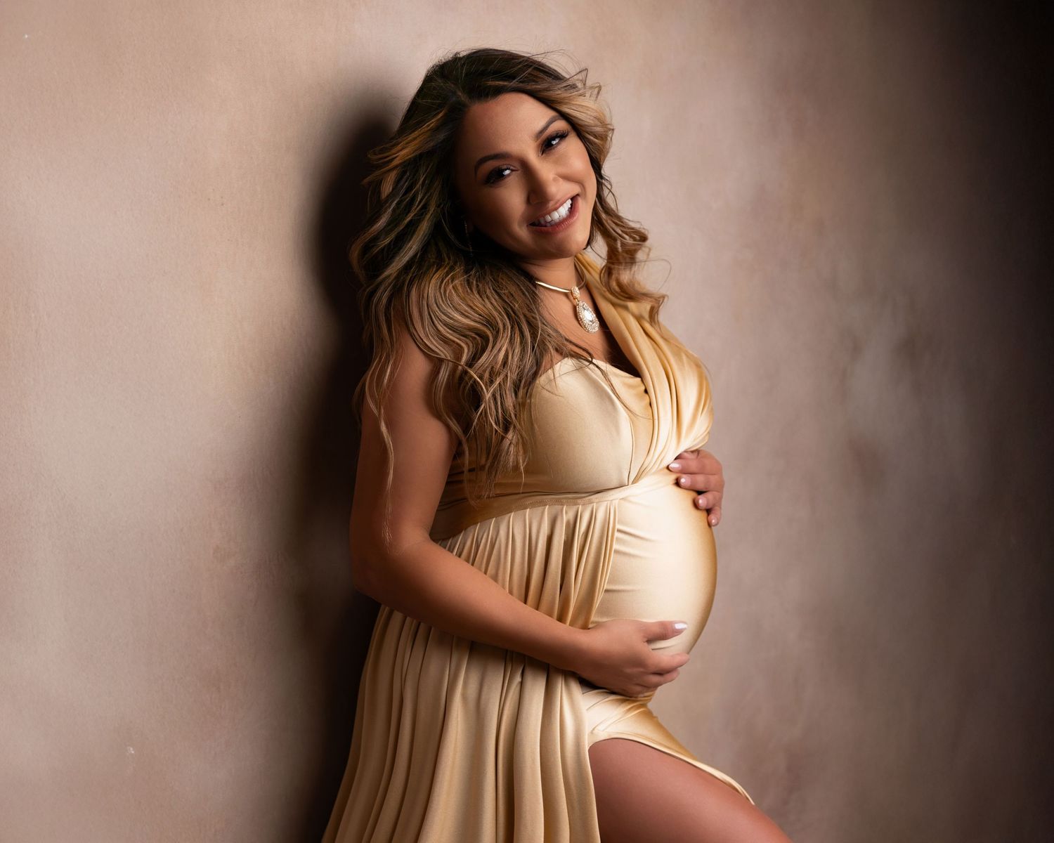 Smiling pregnant woman in elegant gold maternity gown cradling baby bump against neutral studio backdrop.