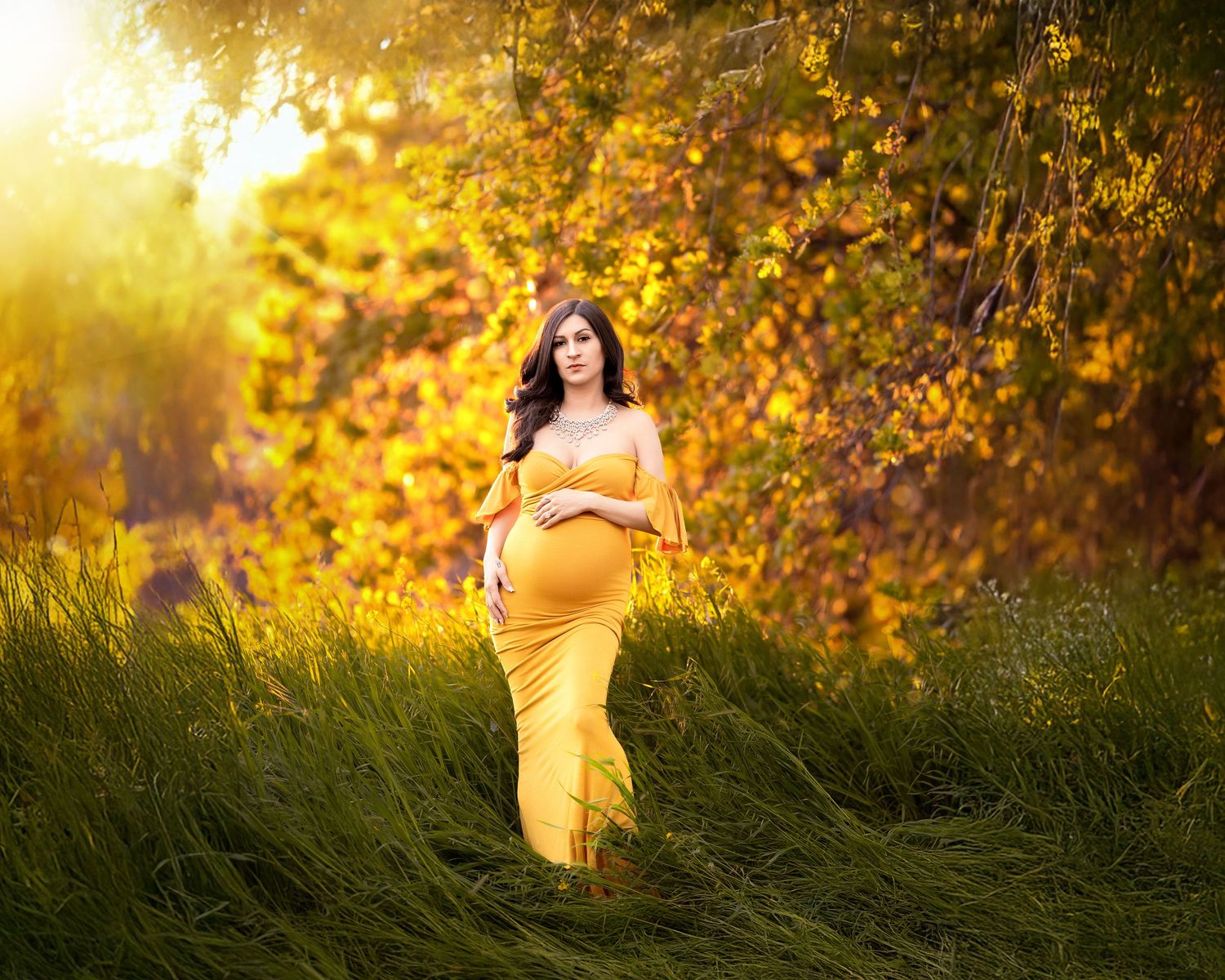 Pregnant woman in yellow gown cradling belly in sunlit autumn meadow.