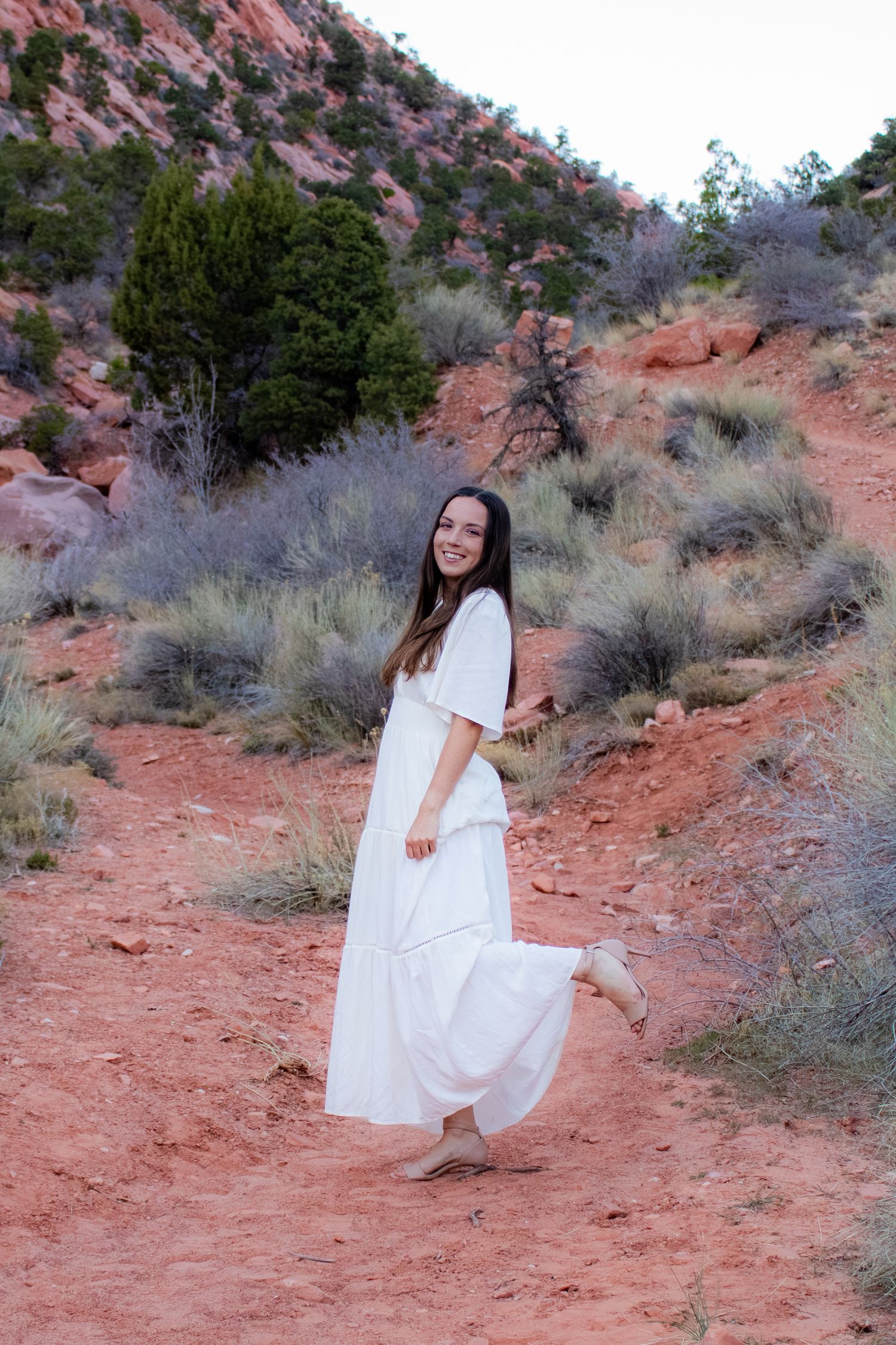 Happy young woman in white dress twirling on red dirt path in desert landscape.