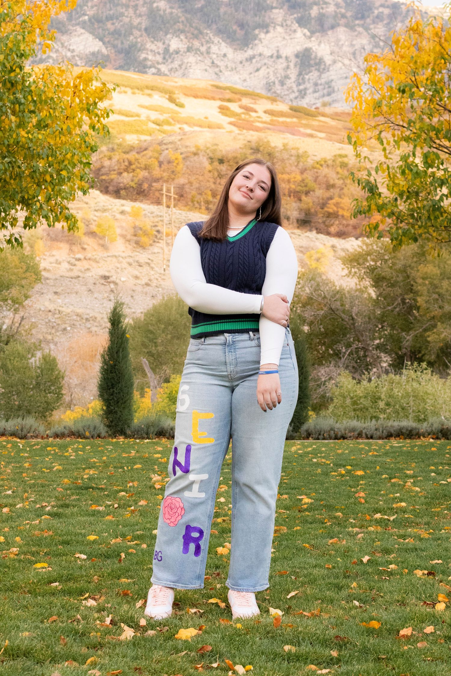 Young woman in navy vest and senior jeans posing in a scenic autumn mountain landscape setting.