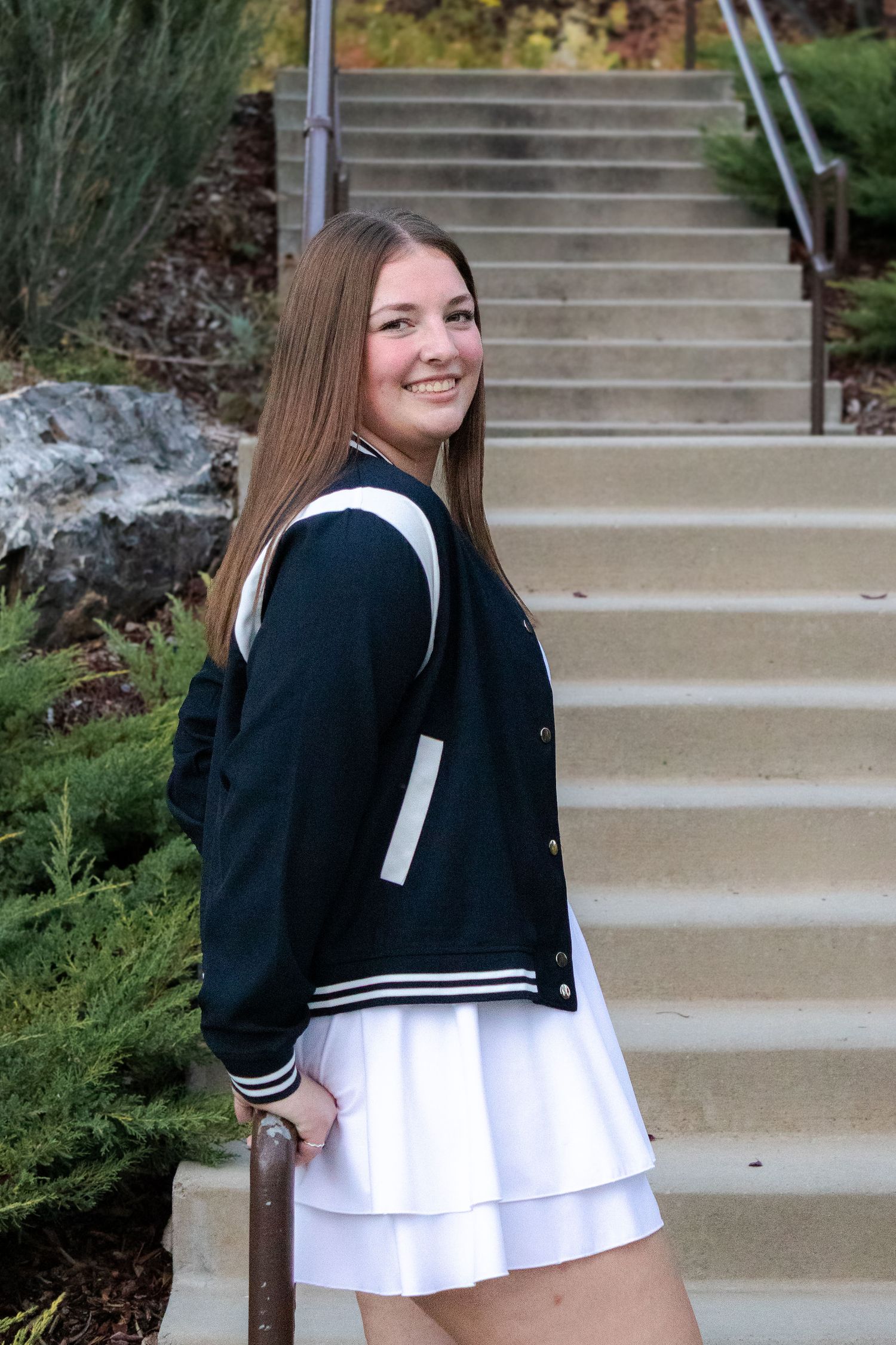 Smiling teen girl in white skirt and varsity jacket posing on outdoor staircase with greenery.